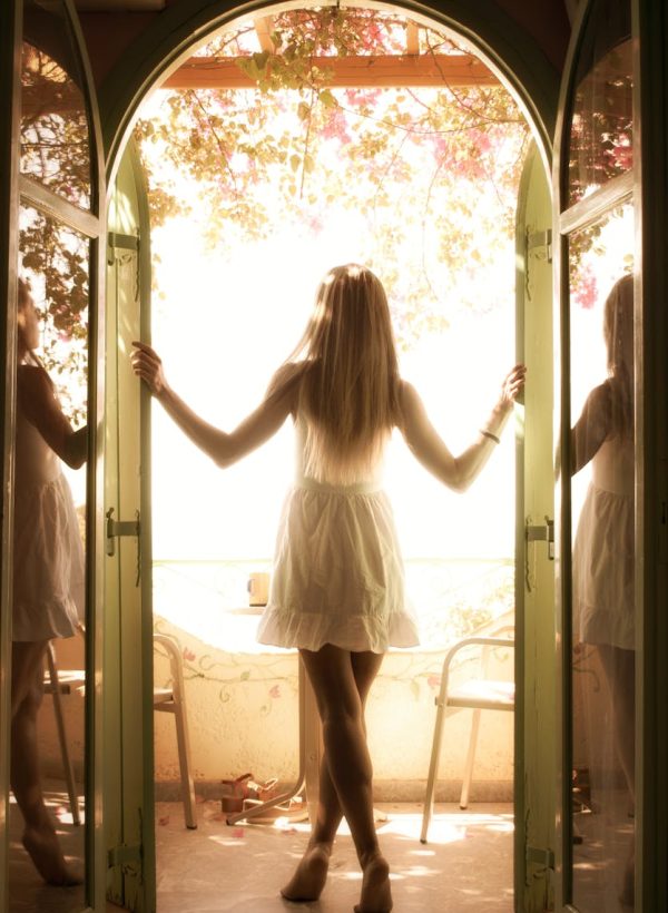 A young woman in a white dress enjoys morning sunlight on a balcony, surrounded by greenery.