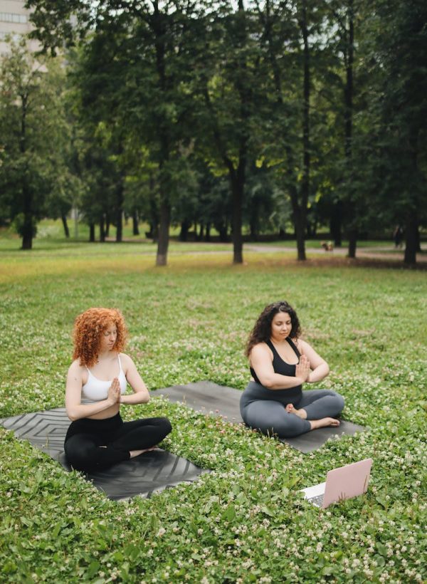 Two women doing yoga on mats in a green park setting, focusing on meditation.